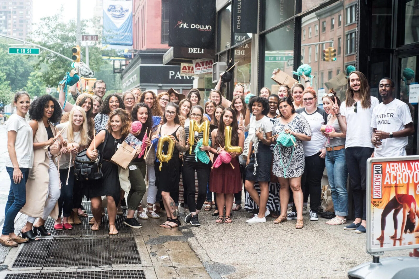 A group of Our Maker Life members smiling together on a New York City sidewalk, several holding knitting needles and yarn, with gold balloons spelling OML.